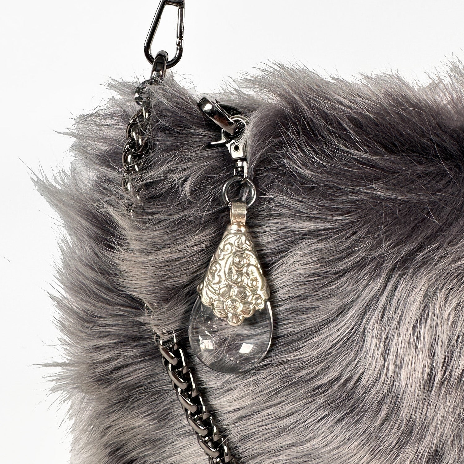 Close-up of the POPPY - GREY LIGHT TIPPED handbag by evokewomen, elegantly crafted with Toscana lamb fur as a crossbody bag featuring a metal chain strap. It showcases a clear teardrop ornament with ornate silver detailing, reminiscent of a Tibetan crystal rock pendant.