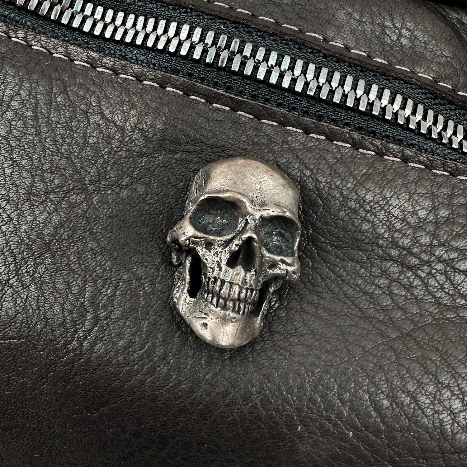 The STELLA - BROWN sling bag by evokewomen features a metallic skull decoration on its textured Italian leather surface, complemented by a sleek, horizontally running zipper above it.