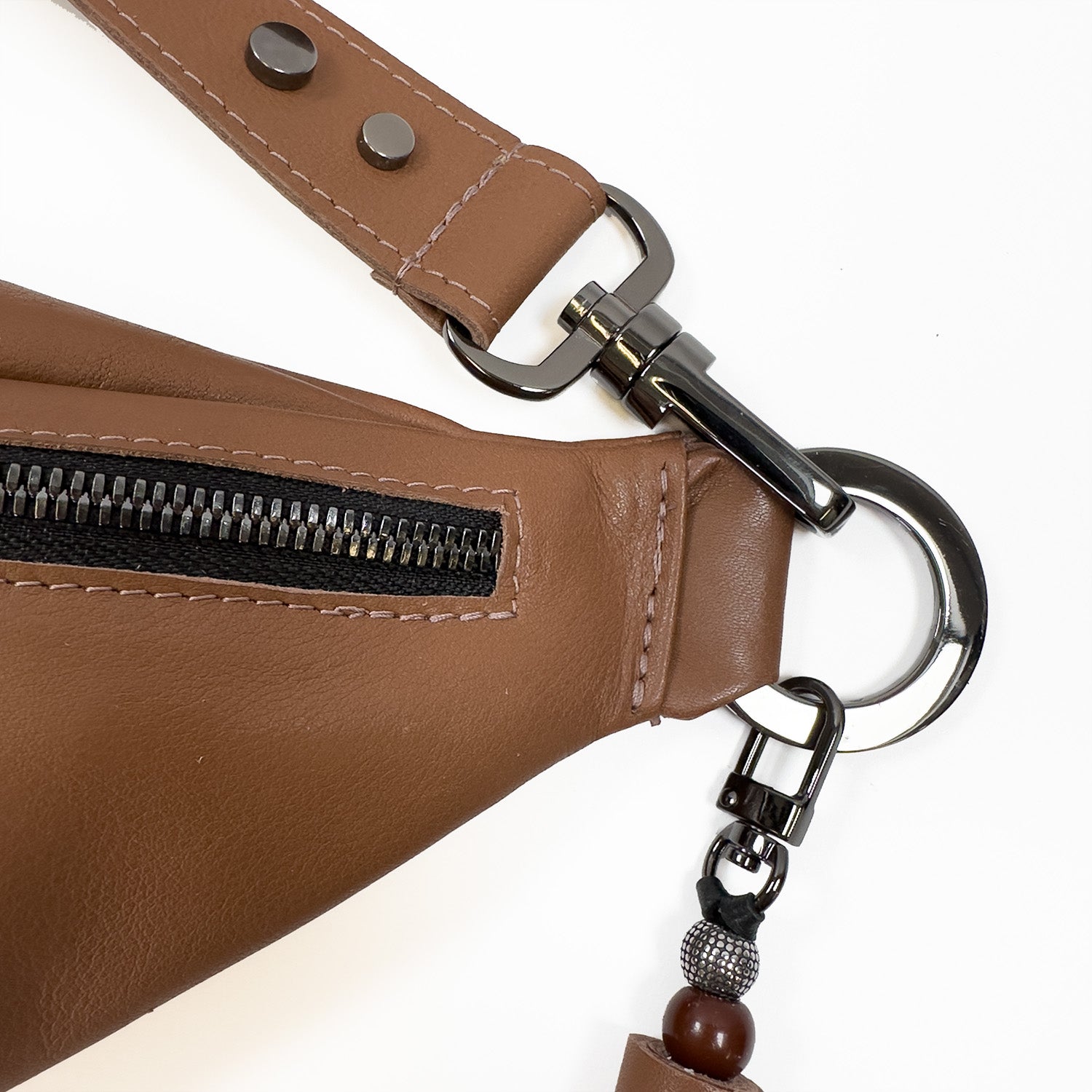 A detailed view of the STELLA - western brown leather crossbody bag by evokewomen, featuring a zipper, a strap with metal buttons, and a keyring decorated with beads.