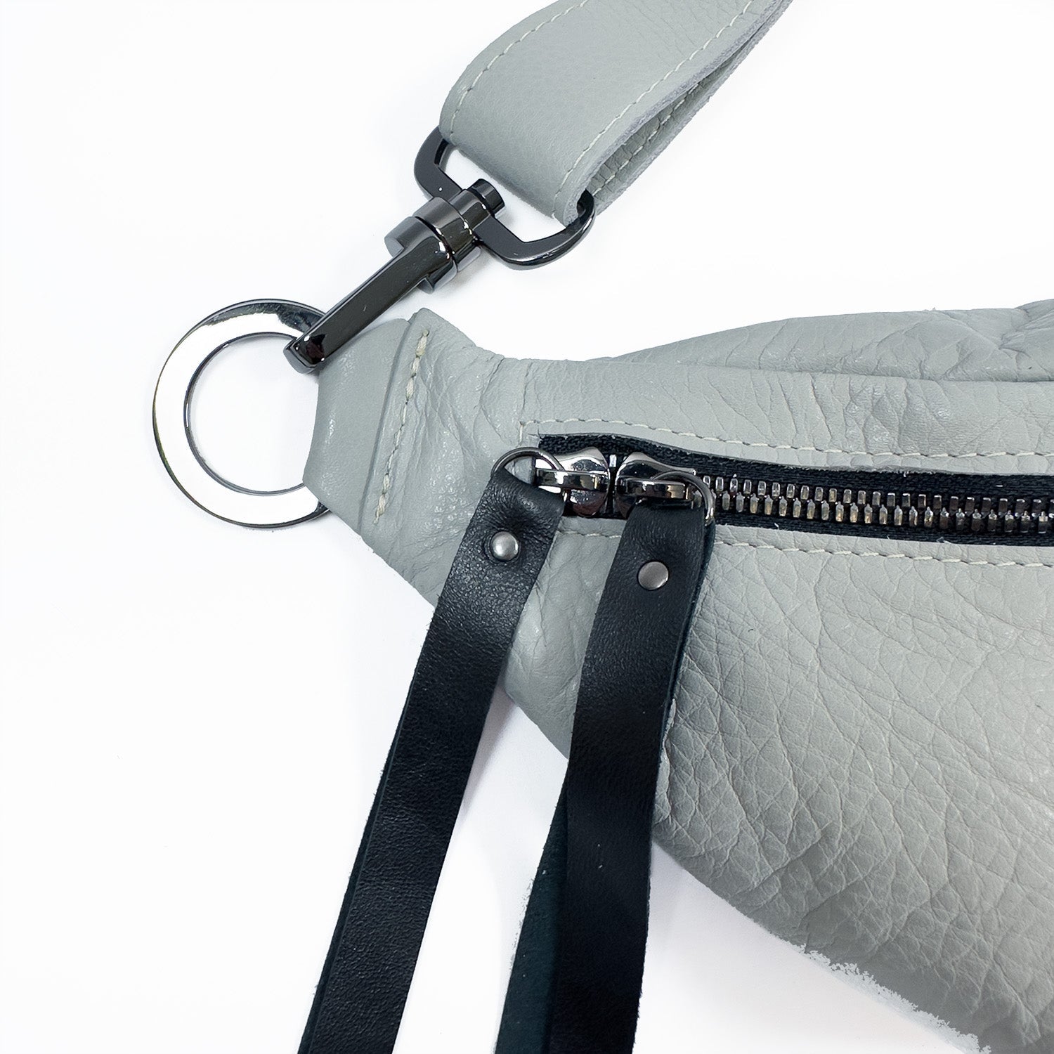 A close-up of the STELLA - light smoke sling bag by evokewomen showcases its gray leather adorned with rivet decorations, a black zipper, a metallic ring, and a black strap attached to a silver clasp.