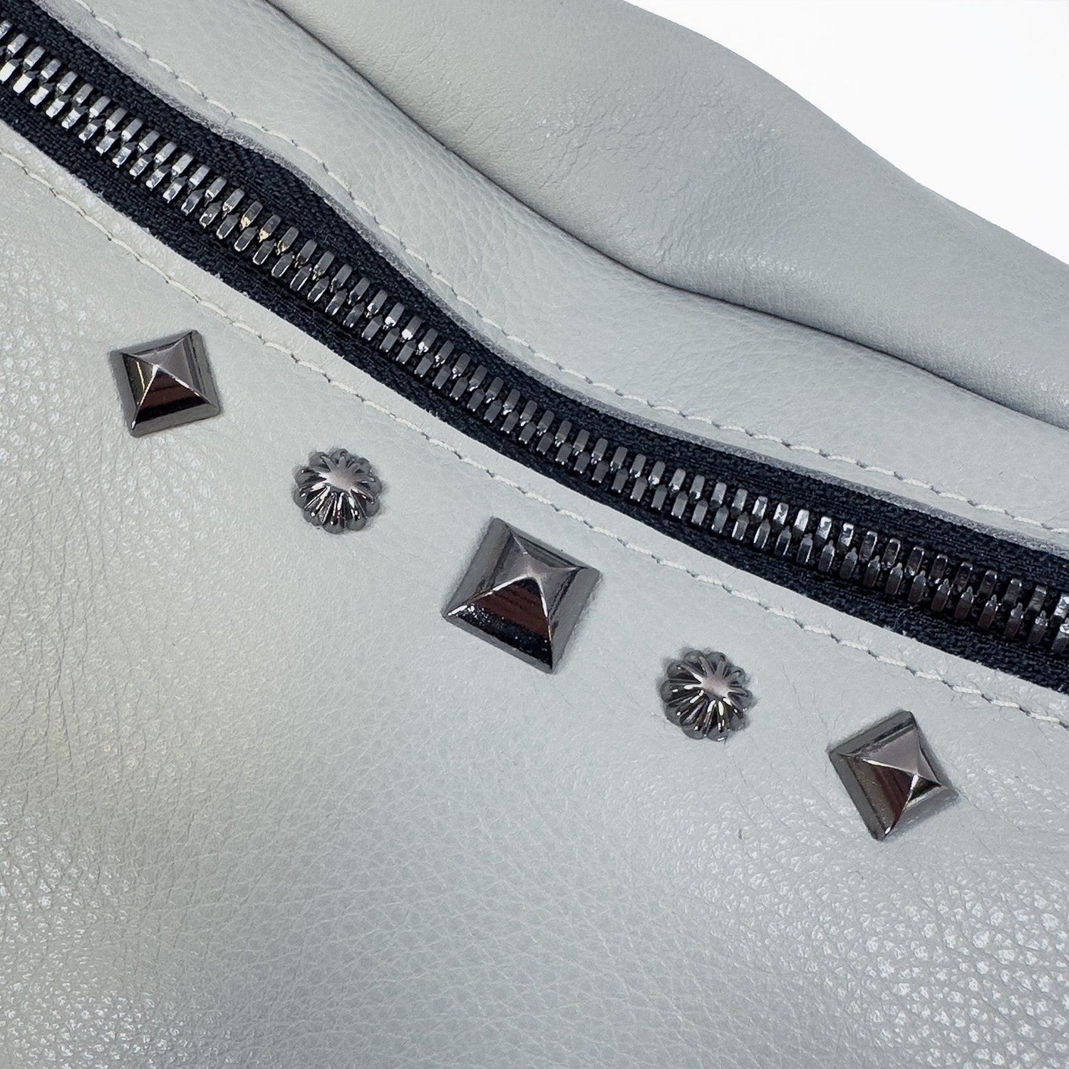 Close-up of the STELLA - light smoke bag by evokewomen, showcasing its gray leather design with a zipper and assorted metallic rivet decorations.