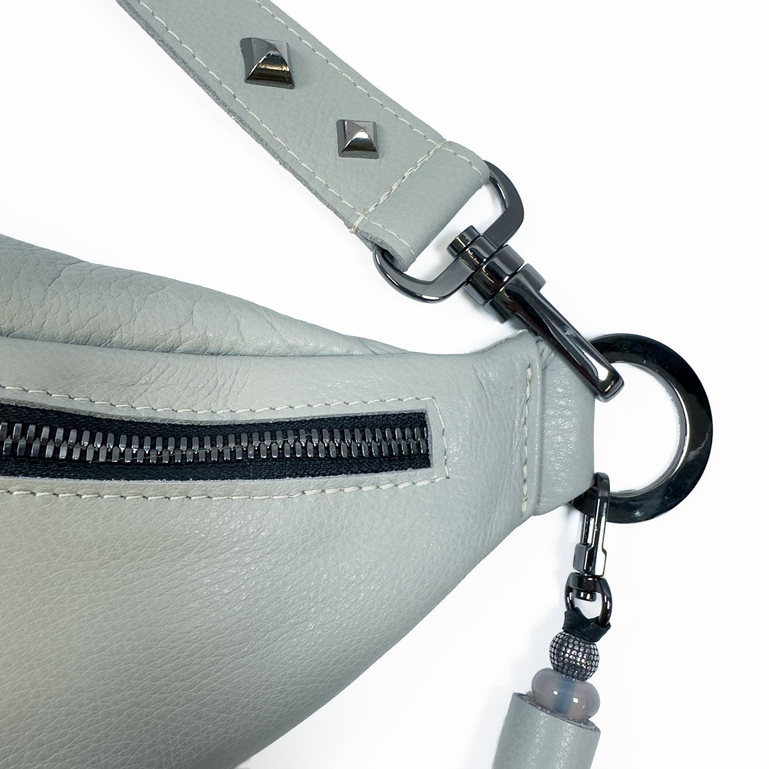 Close-up of the STELLA - light smoke leather sling bag from evokewomen, featuring a black zipper, metallic fixtures, and a decorative beaded tassel attached to a ring. Rivet decorations add an edgy touch to this stylish accessory.