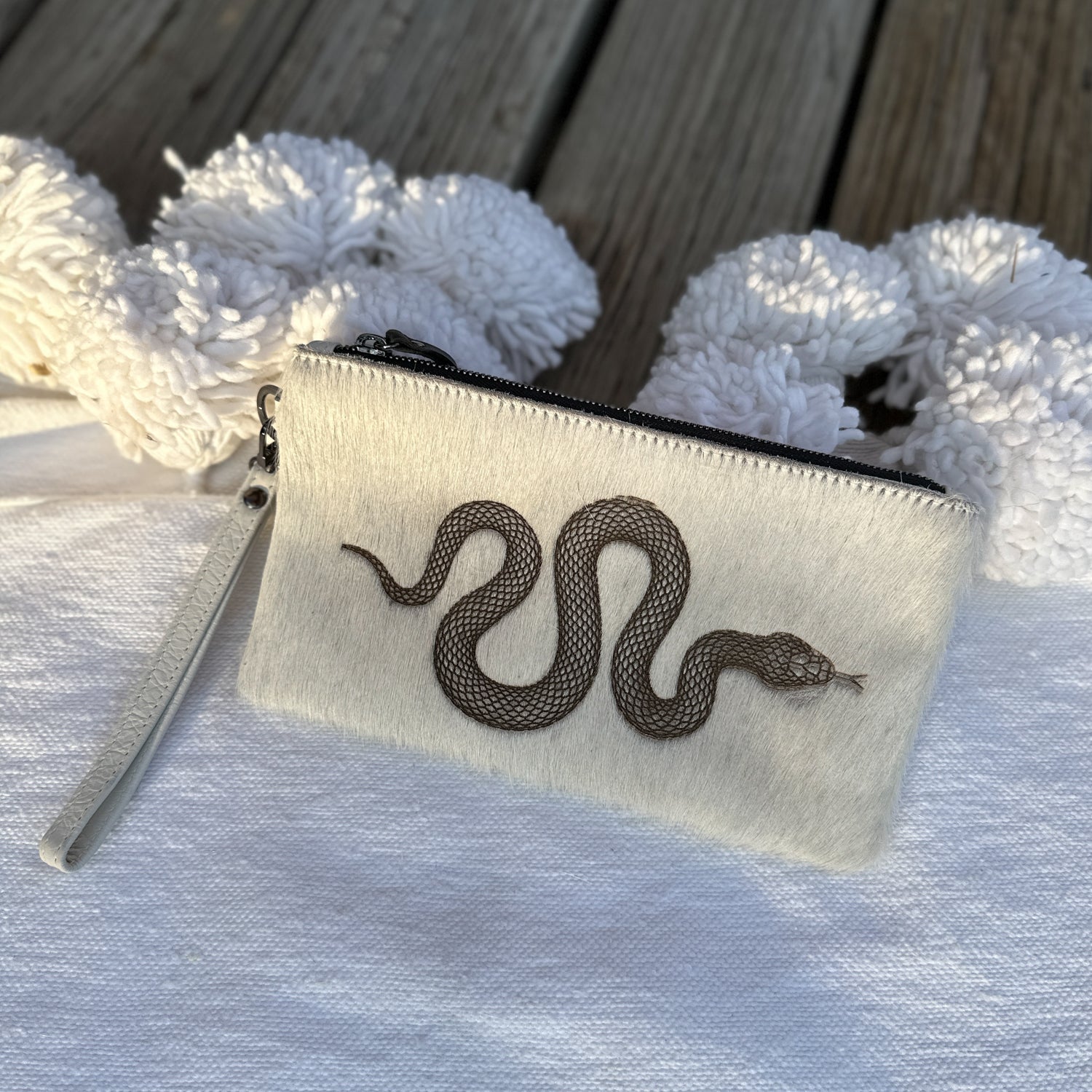 The VICTORIA - off white - laser snake wristlet by evokewomen, featuring a black embroidered snake design, is elegantly displayed on a white surface with wooden flooring in the background.