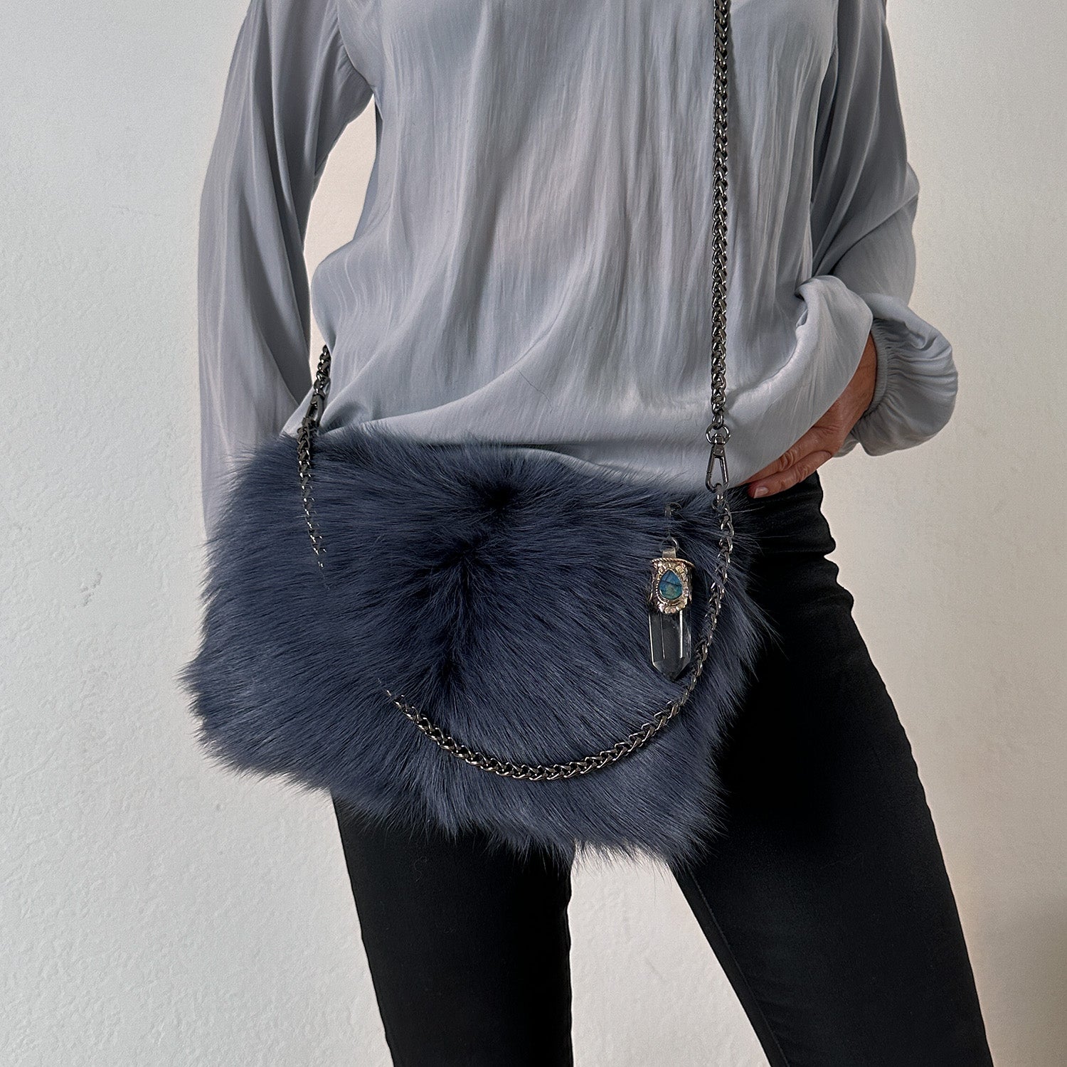 An individual dressed in a gray blouse and black pants is carrying the POPPY - BLUE, a fuzzy navy crossbody bag from evokewomen with a metal chain strap and decorative blue accessory, highlighted by a Tibetan crystal rock pendant.