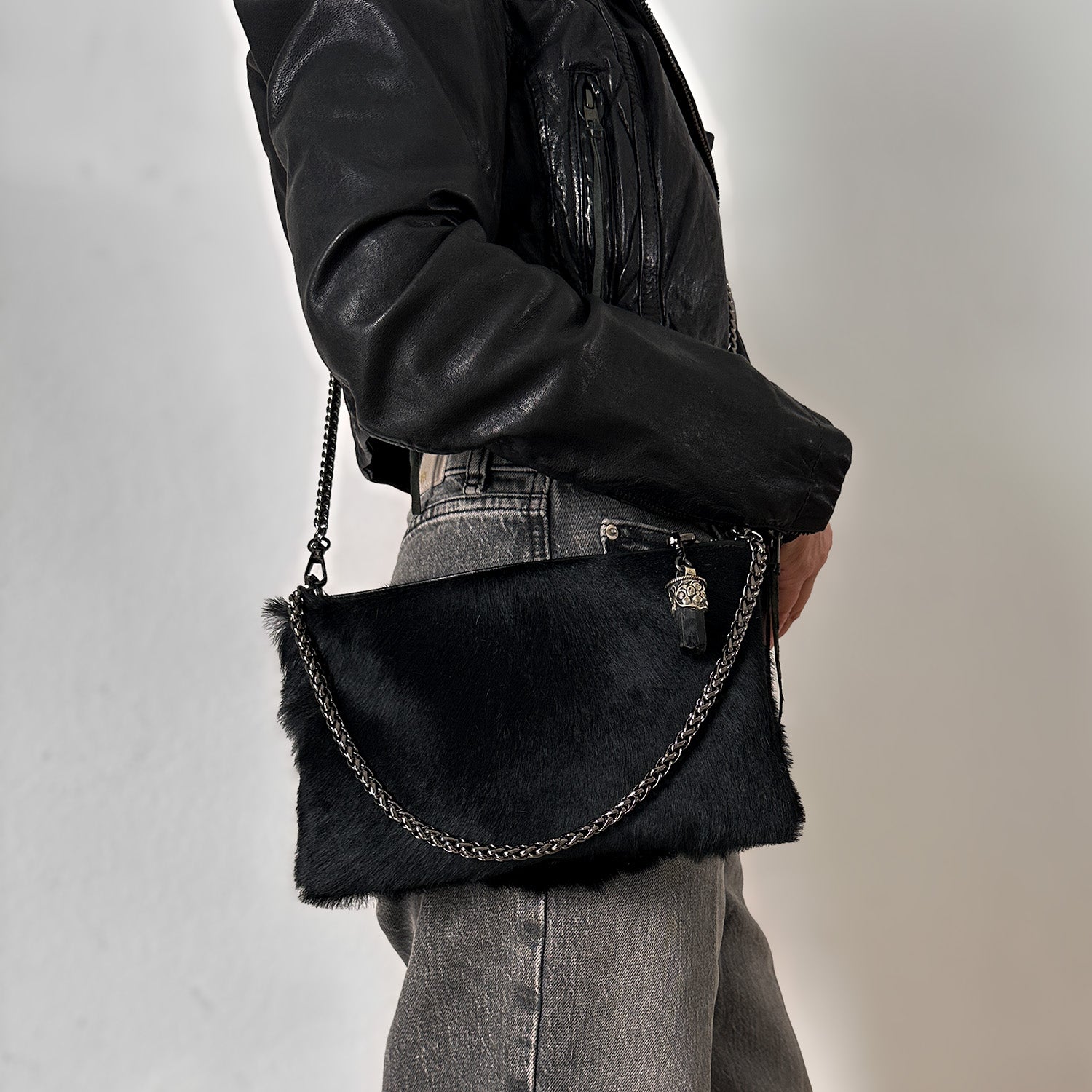 A person wearing a black leather jacket and gray jeans is holding the chic POPPY - BLACK crossbody bag from evokewomen, made from goat fur, with a sleek chain strap elegantly draped over their shoulder.