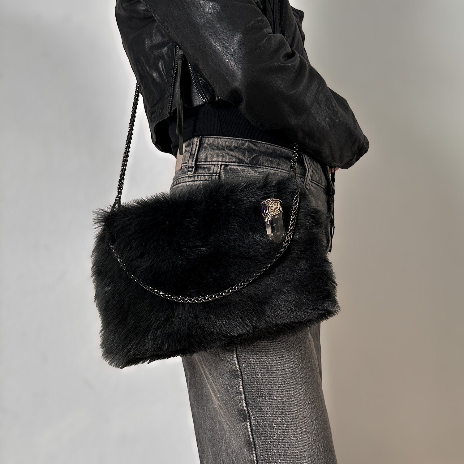 A person dressed in a black leather jacket and gray jeans is carrying the evokewomen POPPY - BLACK LIGHT TIPPED Toscana Lamb Fur crossbody bag with a chain strap at their side.