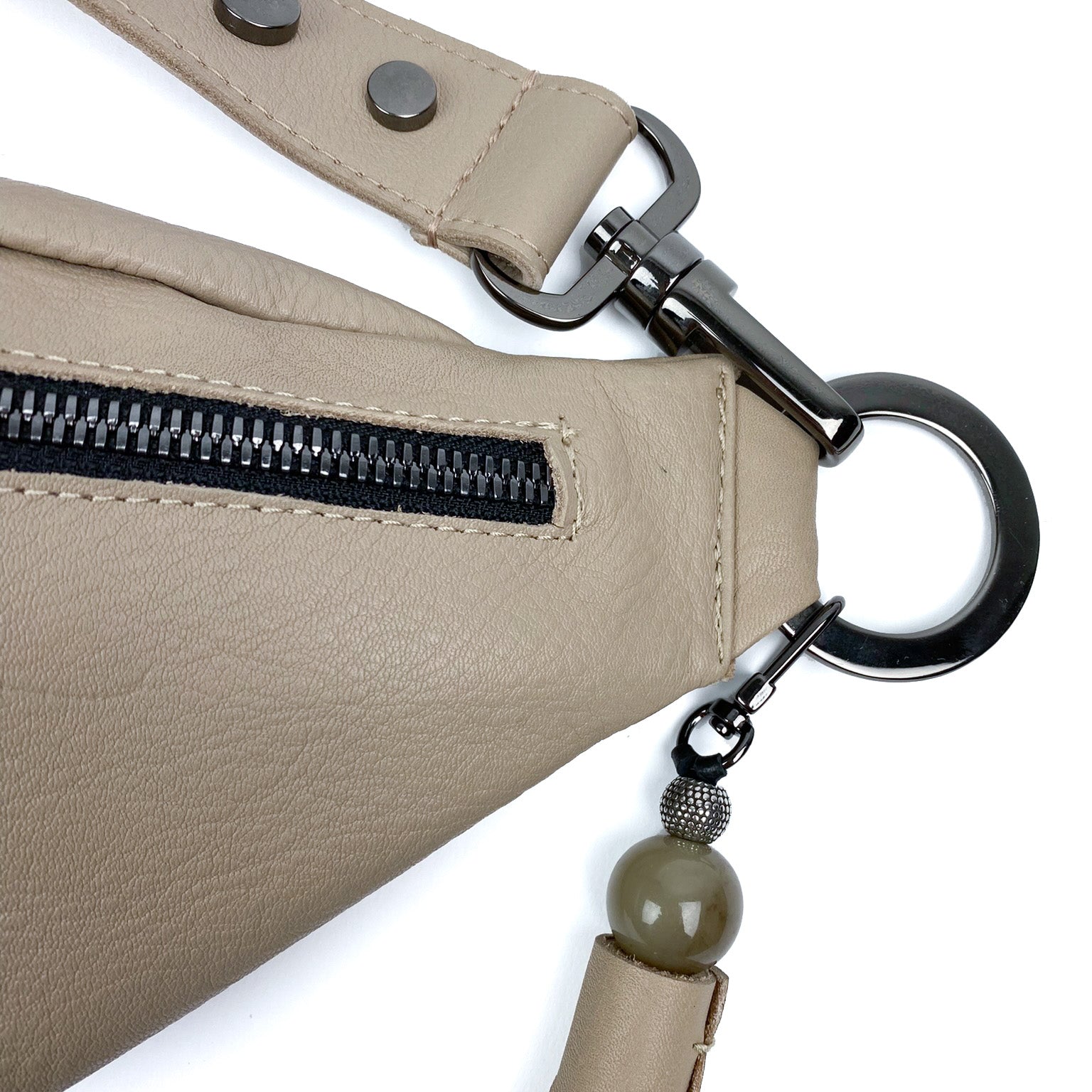 Close-up of the STELLA - SAND sling bag by evokewomen, crafted from beige leather and featuring a black zipper with a large metal ring. The adjustable strap is adorned with a round, olive-colored bead.