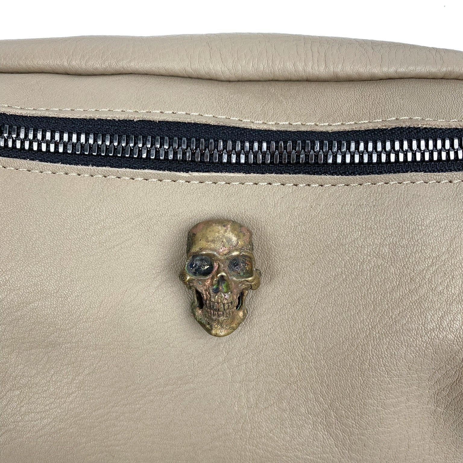 Close-up of the STELLA - SAND beige leather sling bag by evokewomen, featuring a black zipper, an adjustable strap, and a bronze skull decoration.