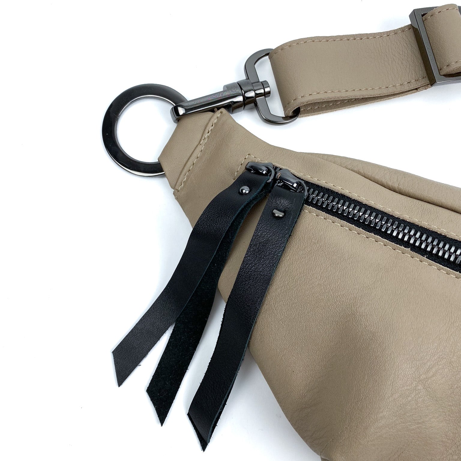 The evokewomen STELLA - SAND is a beige leather crossbody sling bag featuring a black zipper and metal ring attachment, complete with two black leather tassels and an adjustable strap for versatile wear.