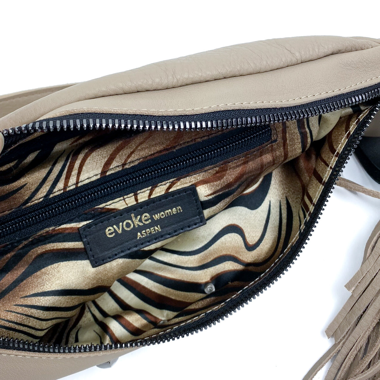 A stylish sand-colored leather sling bag showcases an animal print lining and features a black zipper pocket. Its adjustable strap offers versatility, and the label says "evokewomen STELLA - SAND.