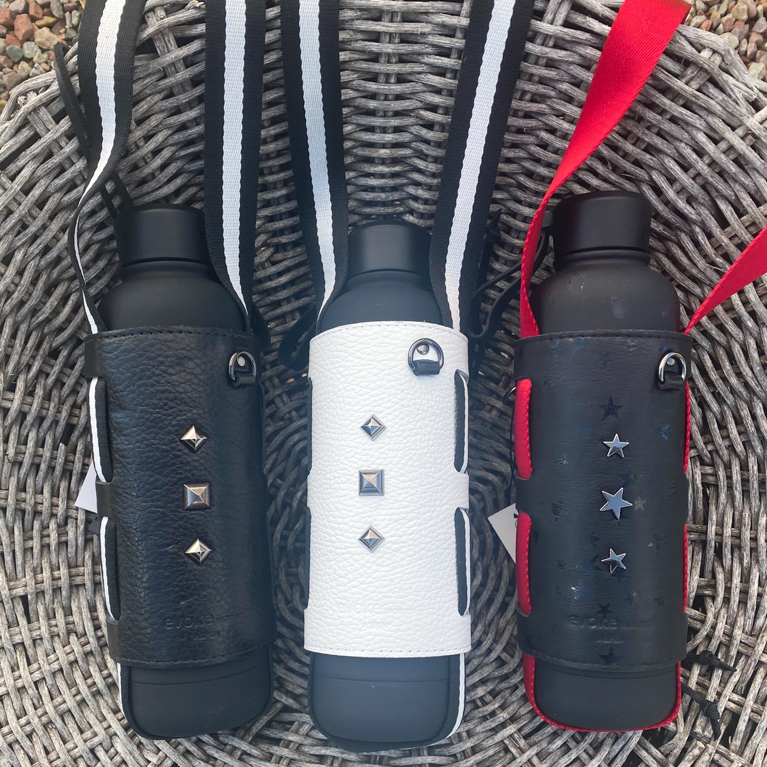 Displayed on a woven surface are three bottles featuring the Carrier - black stars from evokewomen. These water bottles come with star-printed leather covers and adjustable shoulder straps, showcasing unique designs adorned with studs and stars, ideal for fashion-forward bottle carriers.